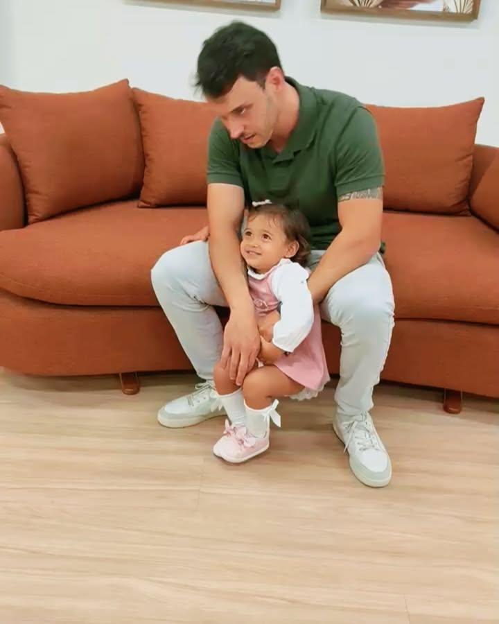 Father lying on a rust sofa lifting his laughing baby daughter overhead