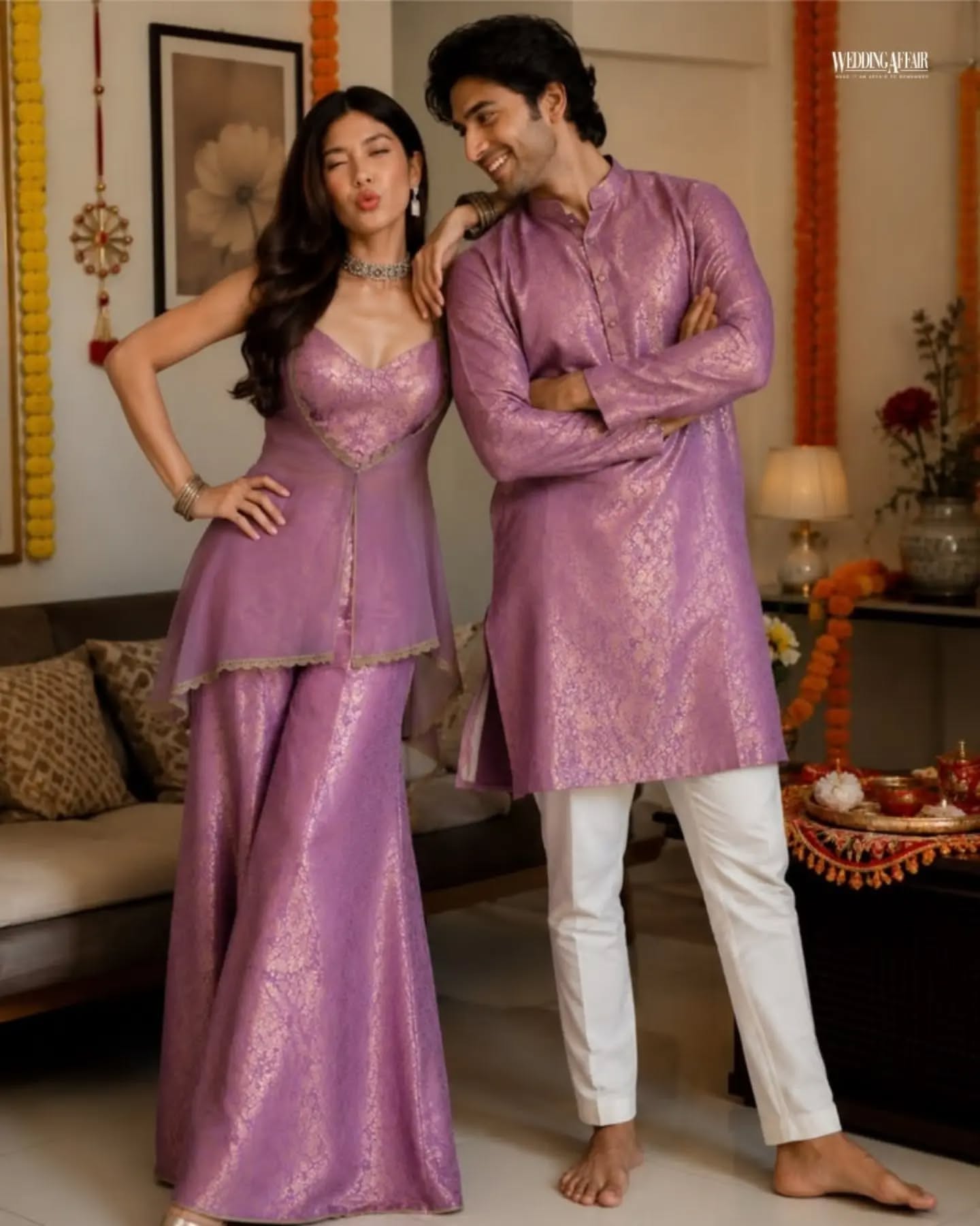 Brother and sister in coordinated pastel ethnic outfits playfully tugging a wrapped gift on a sofa