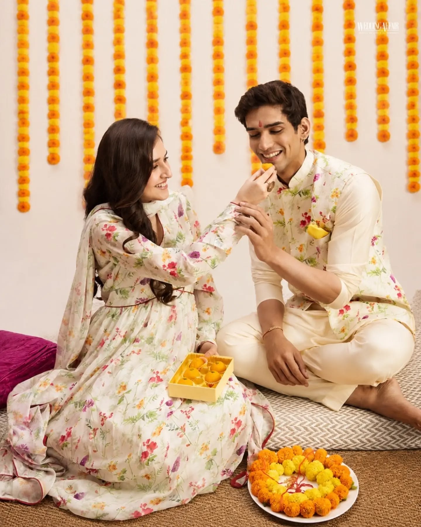 Brother and sister in coordinated pastel ethnic outfits playfully tugging a wrapped gift on a sofa
