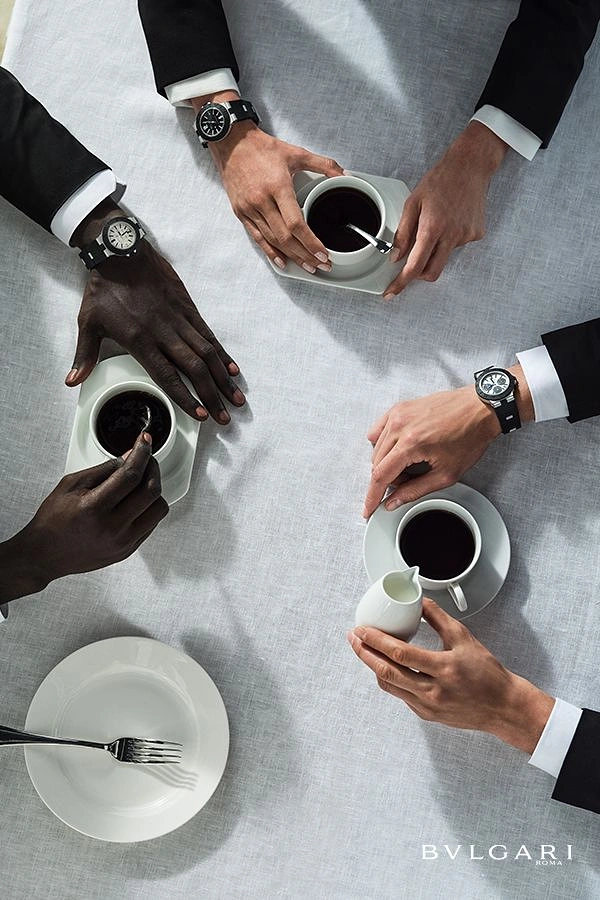 Creative ad: Overhead view of four suited men's hands with sport-luxury watches around coffee cups