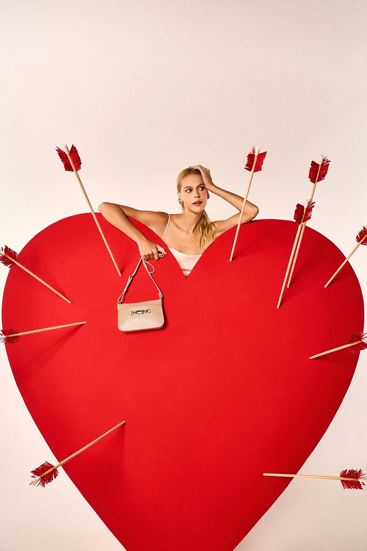 Valentine's Day creative ad: Model leaning over a giant red paper heart pierced by arrows, holding a beige handbag