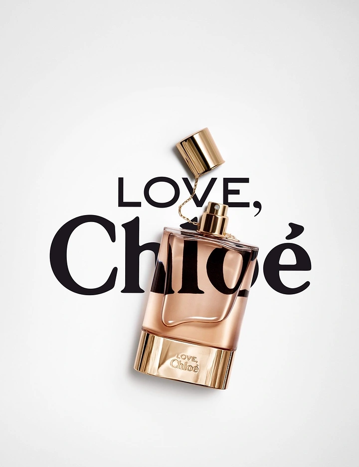 Creative ad: Tilted blush perfume bottle with a gold cap on a chain, over a large wordmark