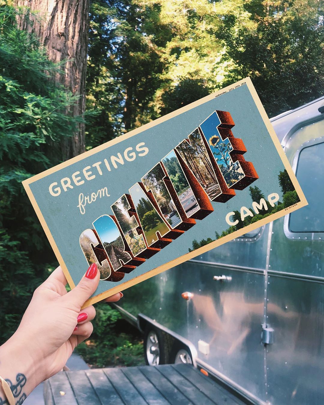 Instagram post: Hand holding a vintage 'Greetings from' large-letter postcard in front of a forest and an Airstream trailer