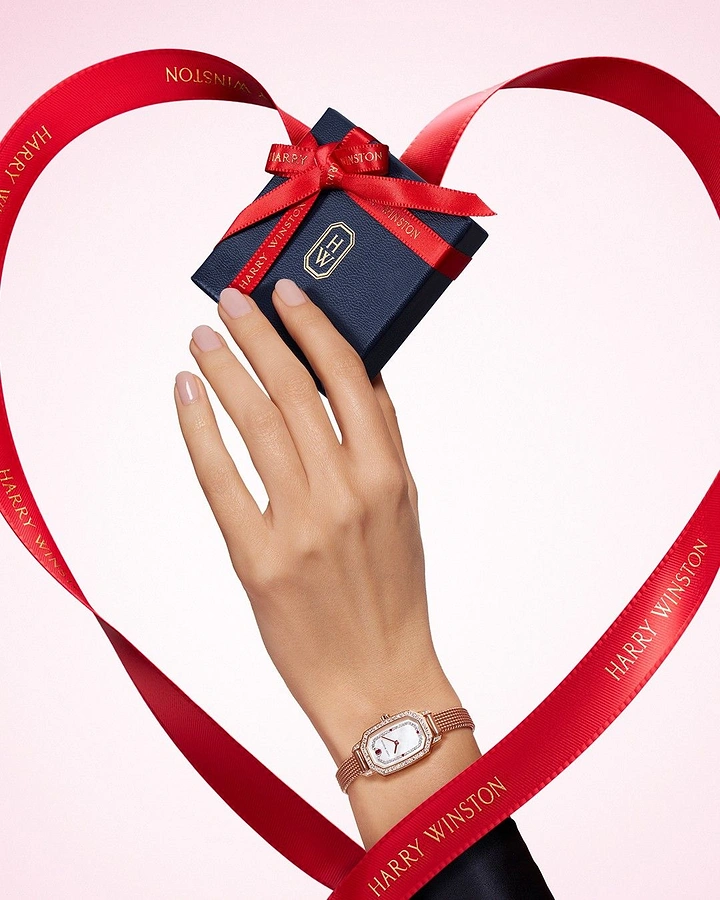 Valentine's Day creative ad: Hand holding navy gift box wearing rose-gold diamond watch, framed by ribbon heart