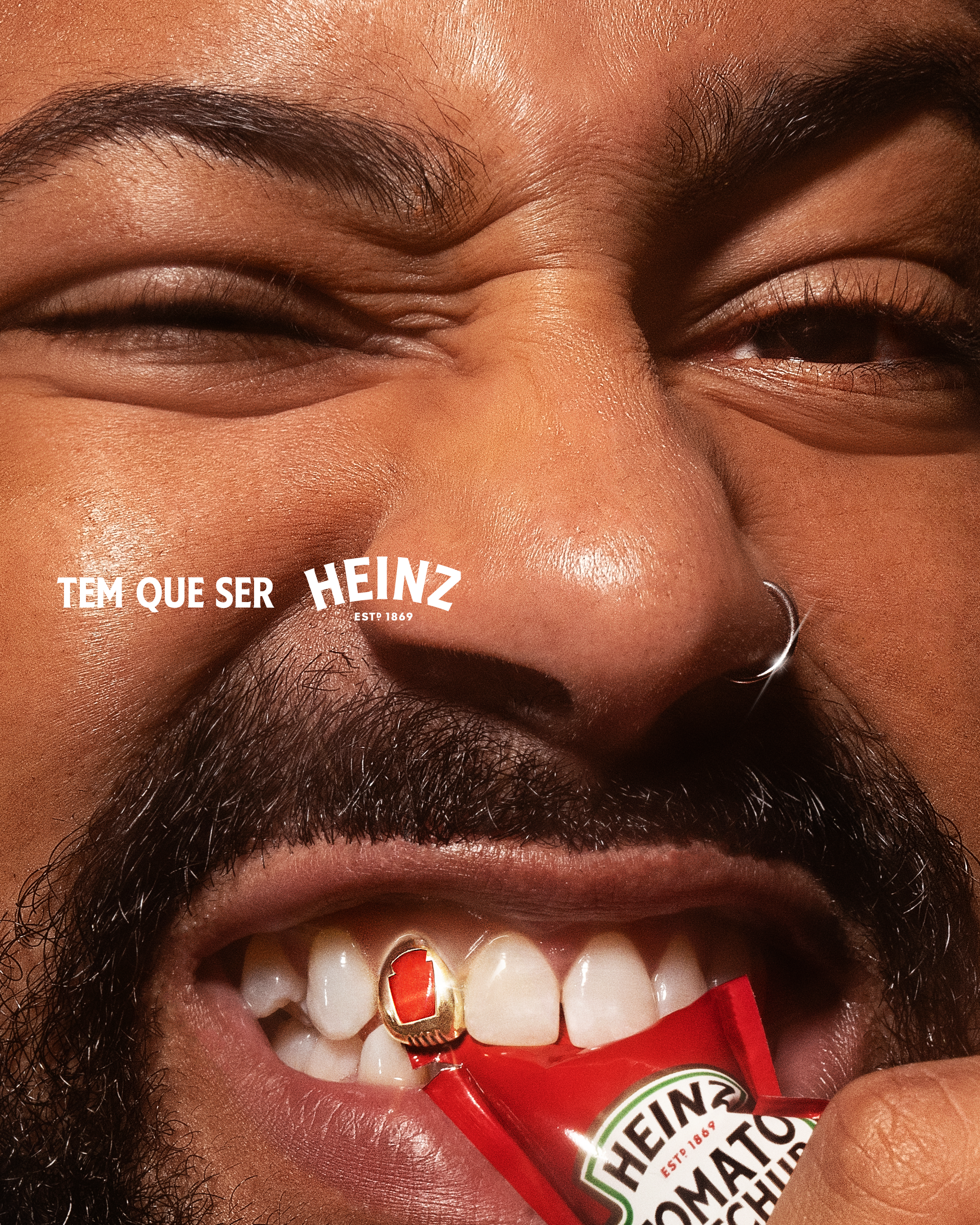 Instagram post: Extreme close-up of a grinning man biting a ketchup sachet, with a gold grill shaped like the brand icon