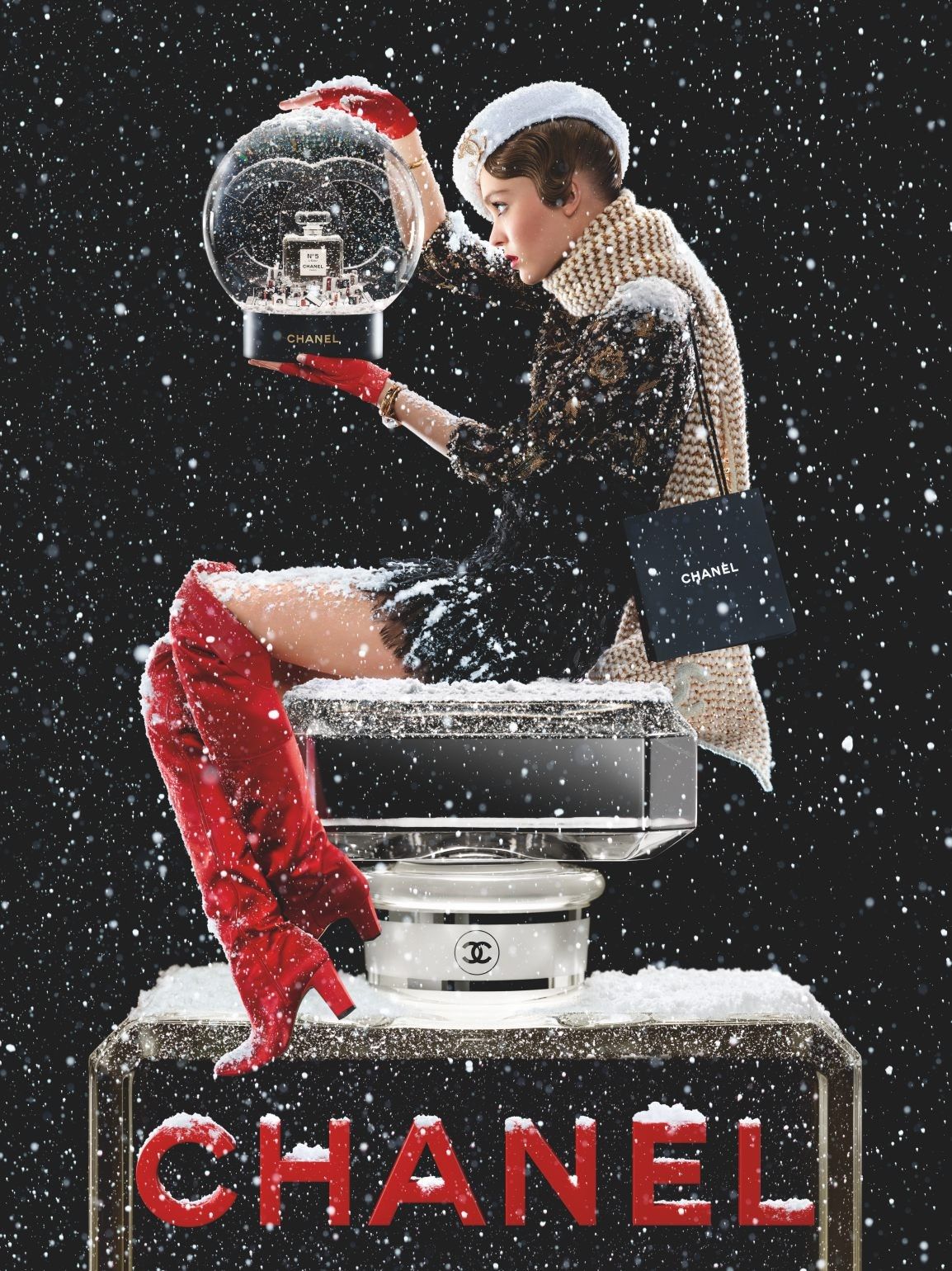 Model perched on a giant perfume bottle holding a snow globe in falling snow