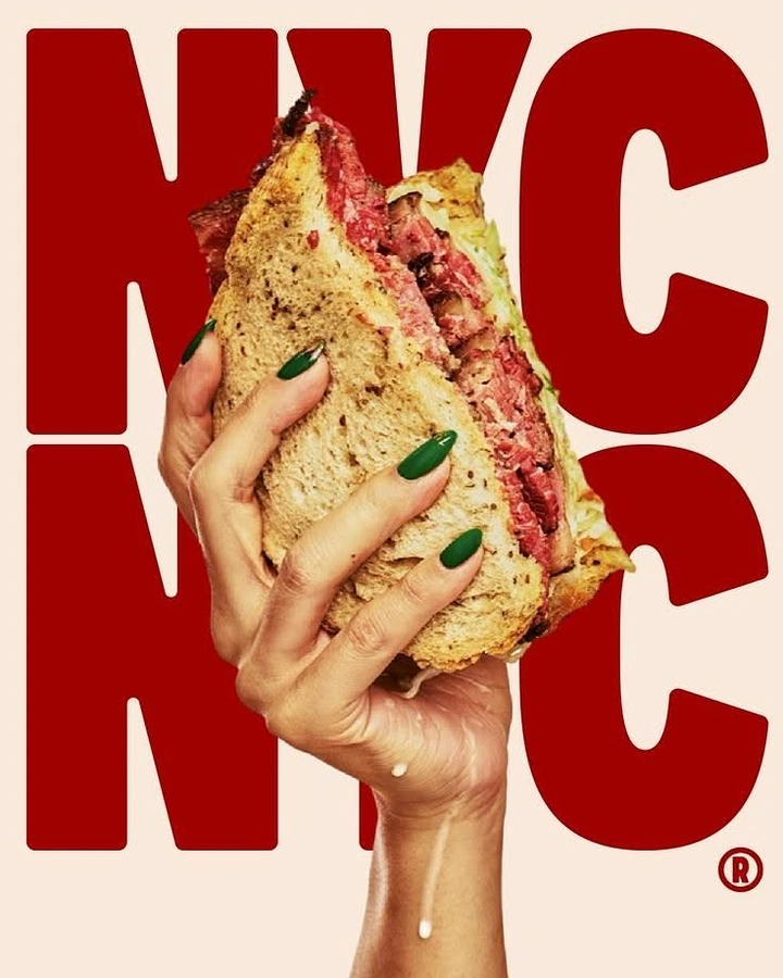 Creative ad: Hand with green nails holding up a thick pastrami rye sandwich